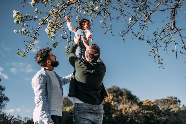 Pareja gay masculina levantando a su joven hija para tocar las flores de un árbol Concepto de familia moderna