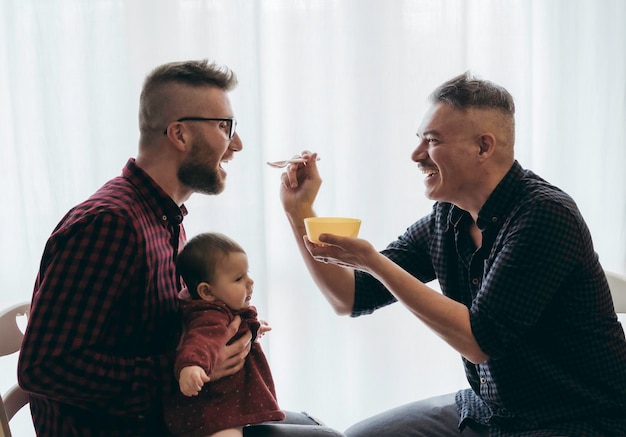 Pareja gay masculina con niña adoptada en casa Dos papás guapos alimentan a la niña en la cocina Niñeros masculinos Familia Lgbt en casa Concepto de diversidad