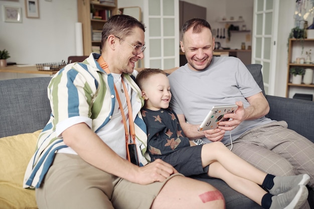 Una pareja gay sentada en un sofá con un hijo pequeño y riéndose mientras ve dibujos animados en una tableta digital