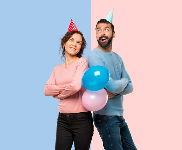 Pareja con globos y sombreros de cumpleaños mirando hacia arriba mientras sonríe sobre fondo rosa y azul