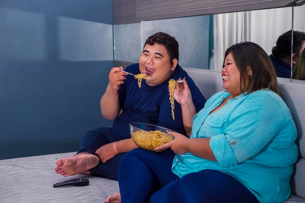 Pareja gorda comiendo fideos felizmente mientras ve la televisión