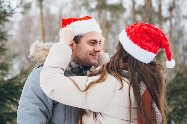 Pareja con un gorro de Papá Noel y abrazos y bromeando en un bosque de coníferas de invierno