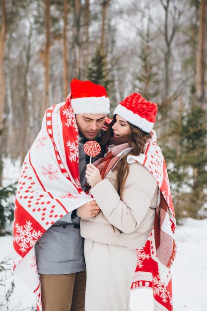 Pareja en un gorro de Papá Noel y envuelto en un plaid abrazos y besos en un bosque de coníferas de invierno