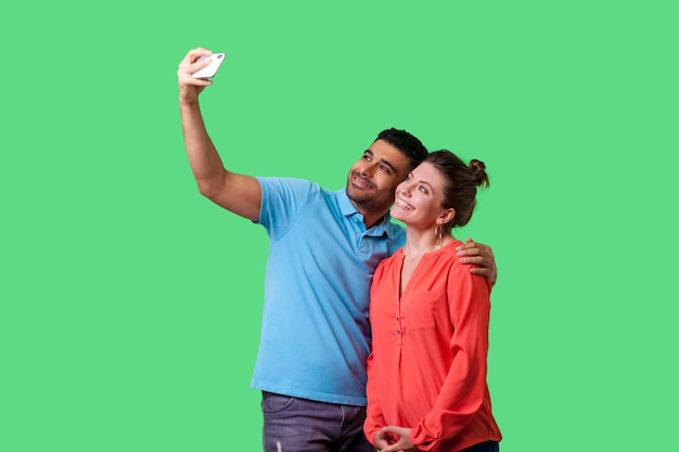 Pareja haciendo selfie Retrato de un joven y una mujer positivos con ropa informal de pie tomando fotos juntos usando recuerdos felices del teléfono celular aislados en una foto de estudio interior de fondo verde