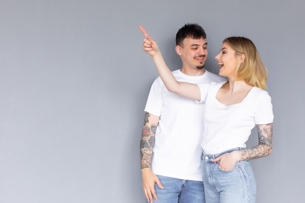 Una pareja hermosa y emocionada con camisetas blancas que se encuentran aisladas sobre un fondo gris apuntando a