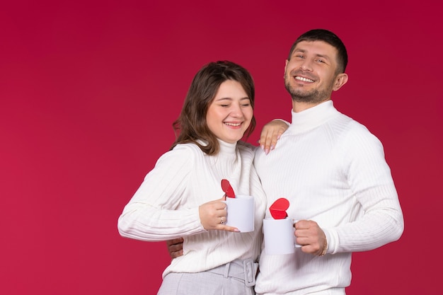 Pareja hermosa con tazas blancas de té y corazones riendo sobre fondo rojo.