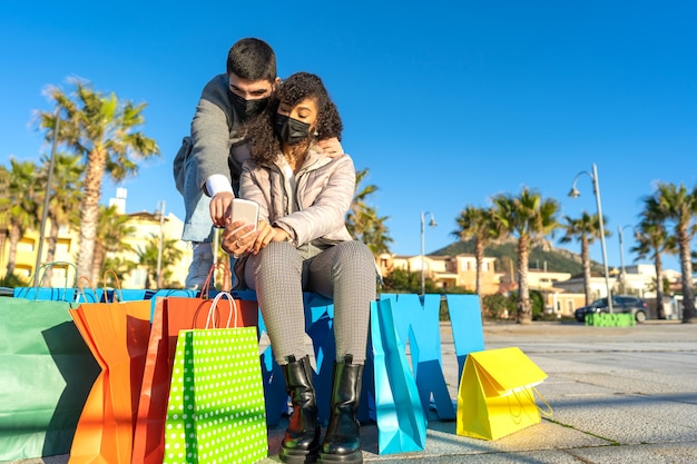 Pareja heterosexual multirracial de jóvenes al aire libre sentados en un banco entre las coloridas bolsas de la compra usando y apuntando el teléfono para comprar en línea con la máscara protectora contra el Coronavirus.