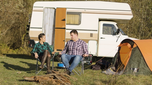 Pareja de hipster sentados en sus asientos de camping y tomados de la mano frente a su autocaravana retro. Carpa para camping.