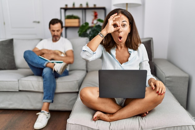 Una pareja hispana de mediana edad en casa una mujer usando una computadora portátil haciendo un gesto de "ok" con un rostro sorprendido un ojo mirando a través de los dedos una expresión de incredulidad