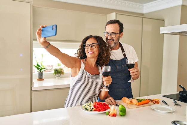 Pareja hispana de mediana edad sonriendo confiada cocinando y haciendo selfie por el teléfono inteligente en la cocina