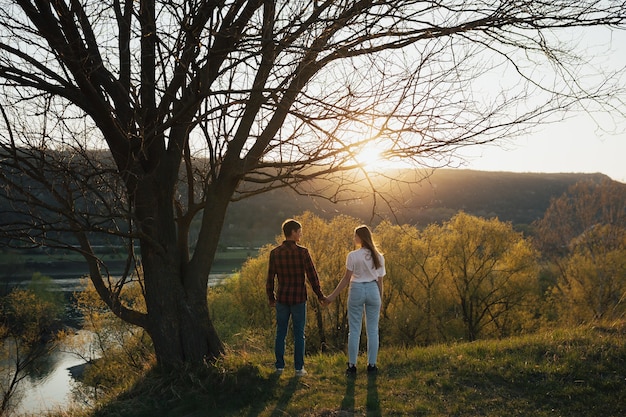 Pareja de hombre y mujer tomados de la mano, mirándose y parados juntos bajo un árbol con la pared del cielo al atardecer.