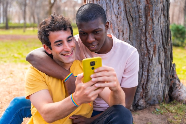 Pareja de hombres multiétnicos en un parque concepto lgbt sentados junto a un árbol divirtiéndose con un móvil amarillo