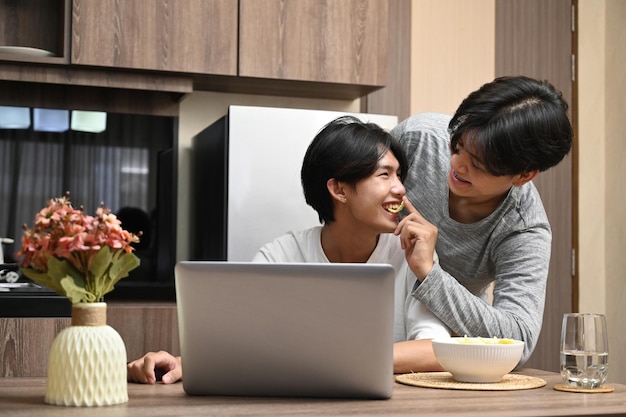 Pareja homosexual teniendo una feliz conversación matutina mientras usa una computadora portátil en la cocina