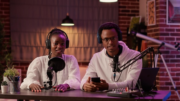 Pareja de influenciadores afroamericanos transmitiendo en vivo vlog dual hablando sentados en el escritorio con la configuración de podcast en el estudio de casa. Creadores de contenido de hombres y mujeres que graban reseñas de teléfonos inteligentes para las redes sociales.