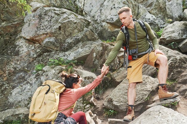 Pareja joven activa con mochilas escalando una colina rocosa juntos hombre sosteniendo la mano de la novia mientras