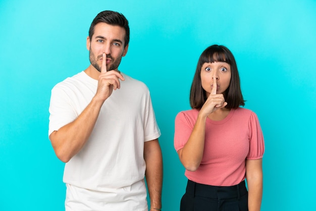 Una pareja joven aislada de fondo azul que muestra un gesto de silencio poniendo el dedo en la boca