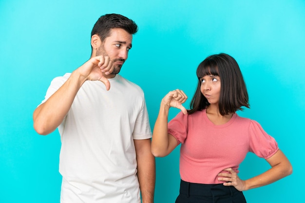 Pareja joven aislada sobre fondo azul mostrando el pulgar hacia abajo con expresión negativa