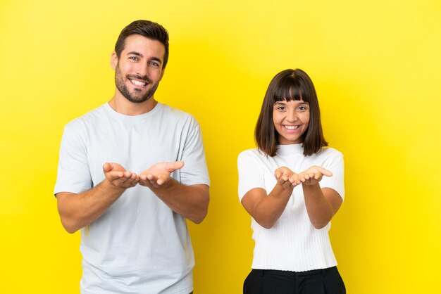 Pareja joven aislado sobre fondo amarillo sosteniendo copyspace imaginario en la palma para insertar un anuncio