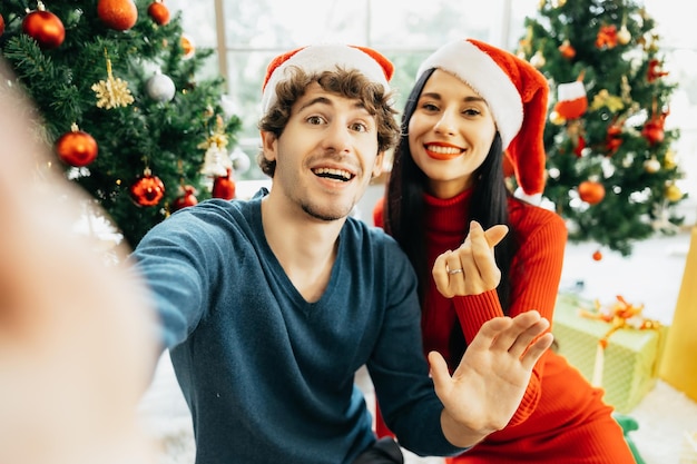 Pareja joven alegre con gorro de Papá Noel en cuarentena sentada rodeada de coloridas cajas de regalo sosteniendo una tableta para videollamada o selfie disfrutando de la celebración de la Navidad en casa.