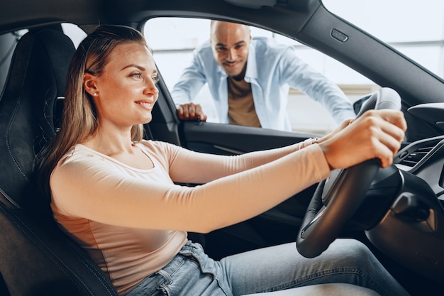 Pareja joven alegre mirando dentro de un coche nuevo que van a comprar en un concesionario de tienda de coches