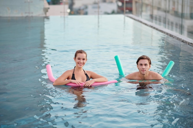 Pareja joven alegre nadando con fideos de piscina en la piscina de la azotea