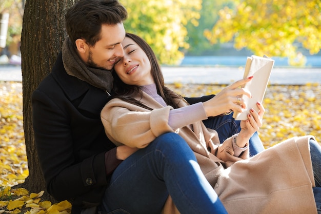 Foto pareja joven alegre pasar tiempo divertido en el parque en otoño, sentado, leyendo un libro