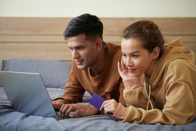 Pareja joven alegre relajándose en la cama y comprando en su tienda en línea favorita disfrutando de la venta de temporada