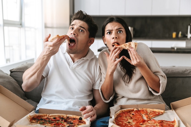 Pareja joven alegre sentado en un sofá en casa, comiendo pizza