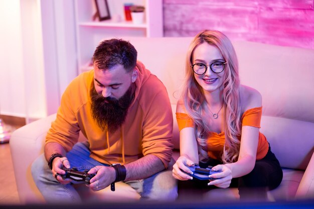 Foto pareja joven alegre sentado en el sofá y jugar videojuegos con controladores inalámbricos. habitación con luz de neón de colores.