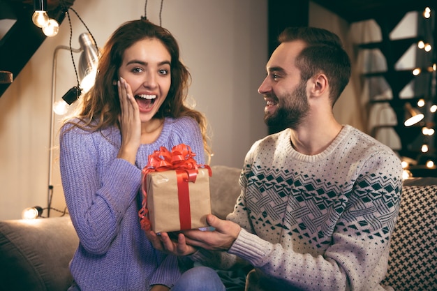 Pareja joven alegre sentados juntos en un sofá en casa, hombre dando un regalo a su novia