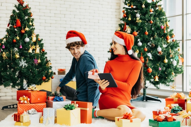 Pareja joven alegre con sombreros de Santa usando tableta sentado rodeado de coloridas cajas de regalo disfrutando de la celebración de la Navidad en casa.