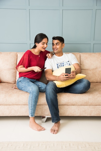 Pareja joven asiática india que usa el teléfono inteligente juntos mientras está sentado en un sofá o sofá y un espacio interior moderno o una casa