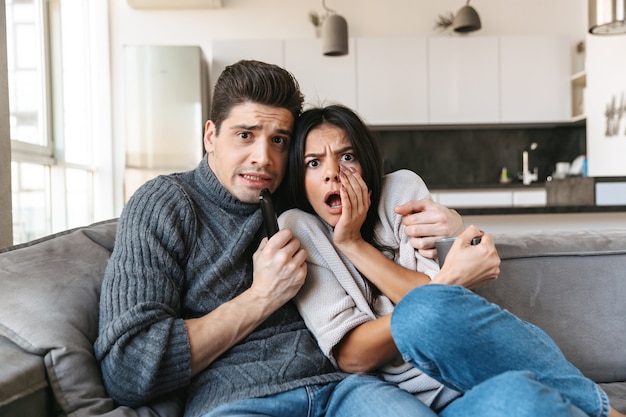 Pareja joven asustada sentada en un sofá en casa, viendo la televisión