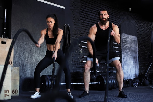 Pareja joven atlética con cuerda de batalla haciendo ejercicio en el gimnasio de entrenamiento funcional.