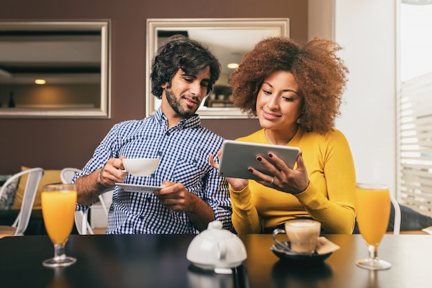 Pareja joven bebiendo un café y un jugo de naranja en la cafetería, usando una tableta