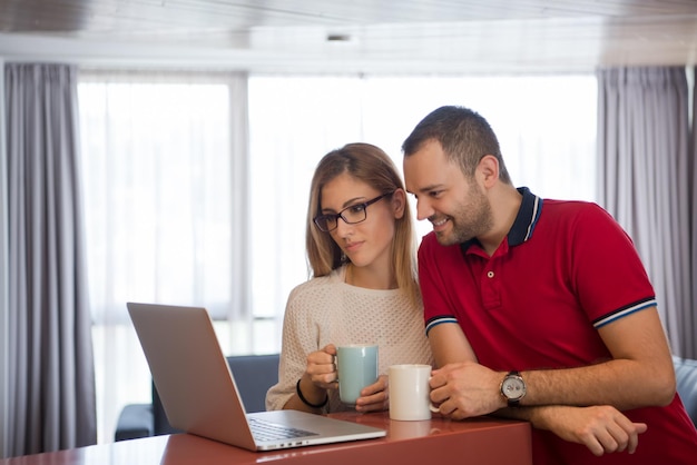 Foto una pareja joven bebiendo café y usando una computadora portátil en una casa de lujo juntos, mirando la pantalla, sonriendo.