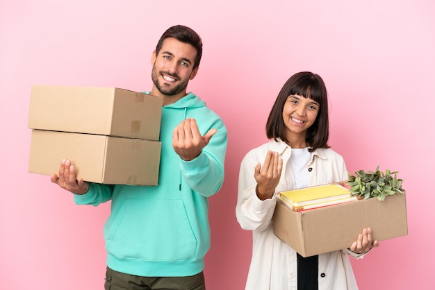 Pareja joven belleza en casa nueva entre cajas aisladas sobre fondo rosa invitando a venir con la mano. Feliz de que hayas venido