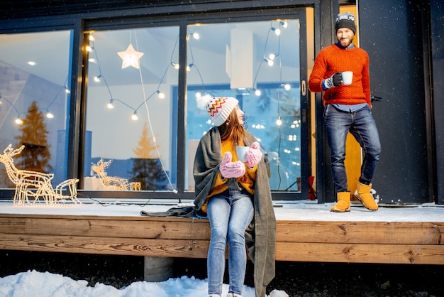 Pareja joven calentando con cuadros y bebidas calientes sentado en la terraza de la casa moderna en las montañas durnig las vacaciones de invierno