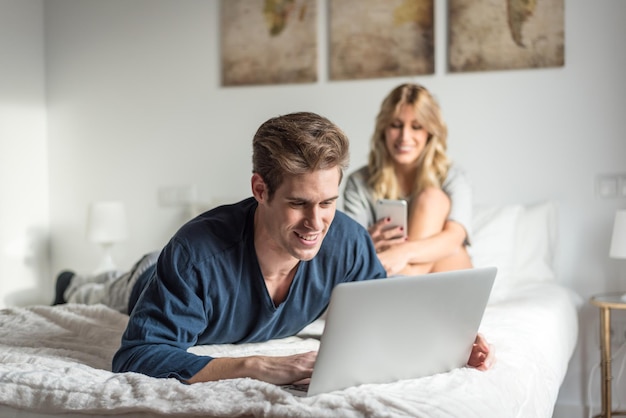 Una pareja joven en la cama entretenida con su computadora y teléfono