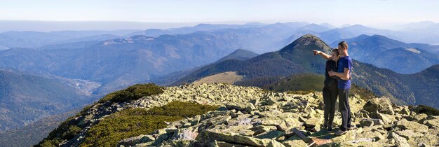 Pareja joven caminata en las montañas de los Cárpatos. Hombre y mujer de pie en la cima de la montaña mirando el hermoso paisaje a continuación. Amplio panorama de altas colinas rocosas en clima cálido de verano. Deportes y actividad.