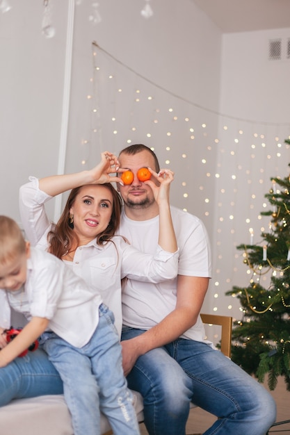 Pareja joven con camisas blancas y jeans divirtiéndose con mandarinas en el sofá contra el árbol de Navidad decorado en casa. Vacaciones de invierno en concepto de hogar acogedor.