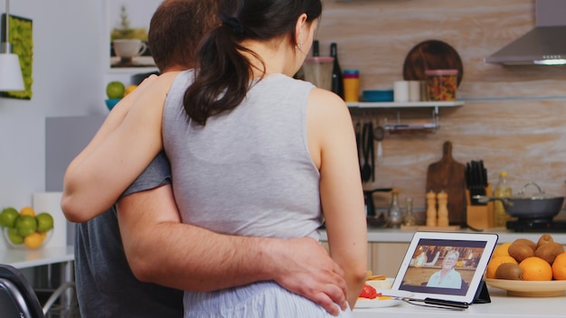 Pareja joven casada en videollamada con madre durante el desayuno en la cocina. Pareja joven en pijama utilizando tecnología en línea web de Internet para charlar con familiares y amigos