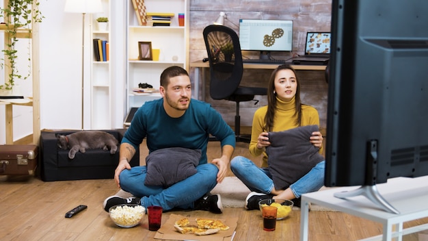 Foto pareja joven caucásica sentados en el suelo en la sala de estar animando a su equipo deportivo favorito. gato durmiendo en el fondo.