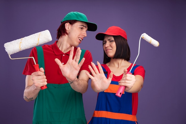 Pareja joven con el ceño fruncido en uniforme de trabajador de la construcción y gorra estirando el rodillo de pintura hacia la cámara haciendo gesto de rechazo chico mirando a chica de lado mirando a cámara aislada en la pared púrpura