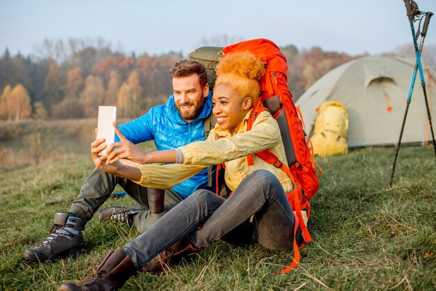 Pareja joven en chaquetas de colores con mochilas divirtiéndose con el teléfono inteligente sentado en el césped cerca del lugar de acampada