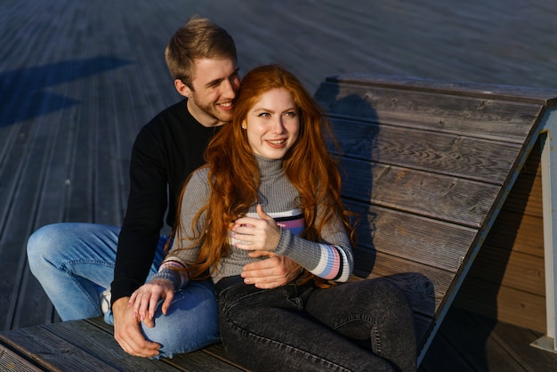 Pareja joven de chico y chica con cabello rojo de apariencia caucásica en ropa casual en un día soleado sentado en el parque en un banco de madera en un abrazo feliz relación entre un hombre y una mujer