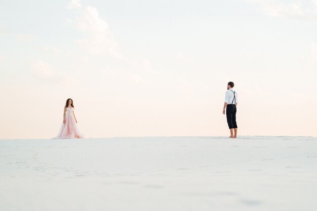 Pareja joven un chico con pantalones negros y una chica con un vestido rosa están caminando por la arena blanca del desierto