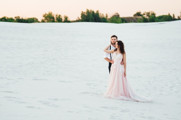 Pareja joven un chico con pantalones negros y una chica con un vestido rosa están caminando por la arena blanca del desierto