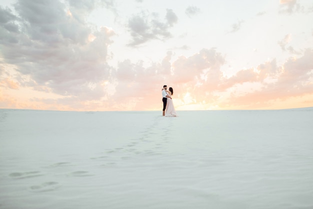 Pareja joven un chico con pantalones negros y una chica con un vestido rosa están caminando por la arena blanca del desierto