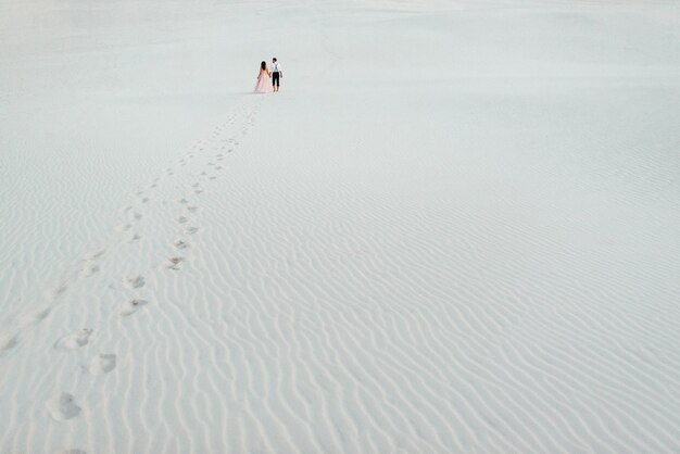 Pareja joven un chico con pantalones negros y una chica con un vestido rosa están caminando por la arena blanca del desierto